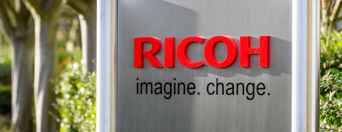 the words Ricoh in red and the words imagine and change in black on silver sign in front of green trees and bushes 