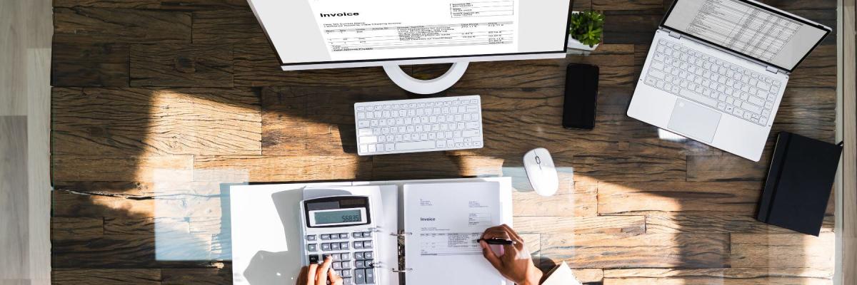 birds eye view of person working on calculator, paper invoices, and computer screens