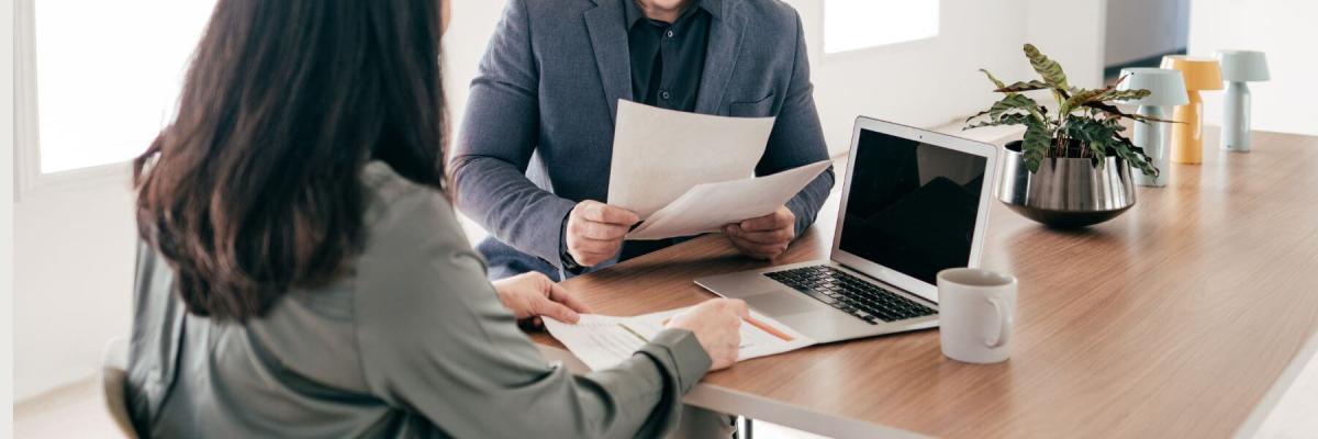 two people discussing documents at a table with laptop